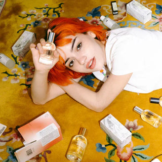 Person with orange hair lies on yellow floral rug surrounded by perfume bottles and DedCool product packaging.